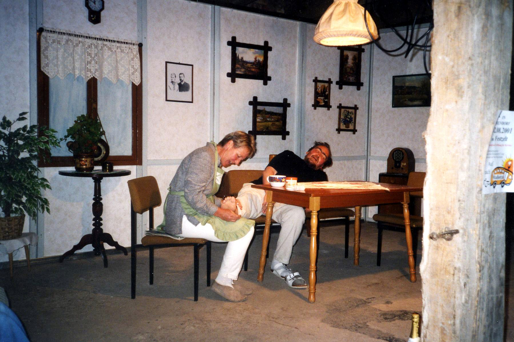 Ein Szenenfoto aus der Theatersaison 2001: Eine Frau und ein Mann sitzen an einem Tisch. Die Frau inspiziert den Fuß des Mannes, den sie auf ihren Knien liegen hat. Der Mann scheint unter Schmerzen zu leiden.